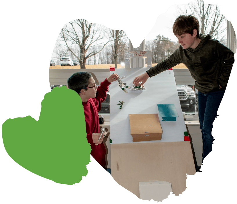 Two children are playing with a toy car on a white ramp indoors. One child is placing the car while the other watches. Trees and cars can be seen outside through the window. There is a graphic of a green heart to the left.