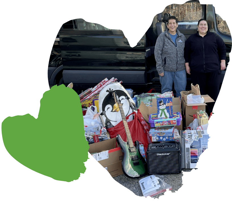 Two people stand next to a car filled with donations, including toys, a large stuffed penguin, a guitar, and an amplifier. Bags and boxes filled with various items are stacked on the ground in front of them. Graphic of a green heart is set to the left.