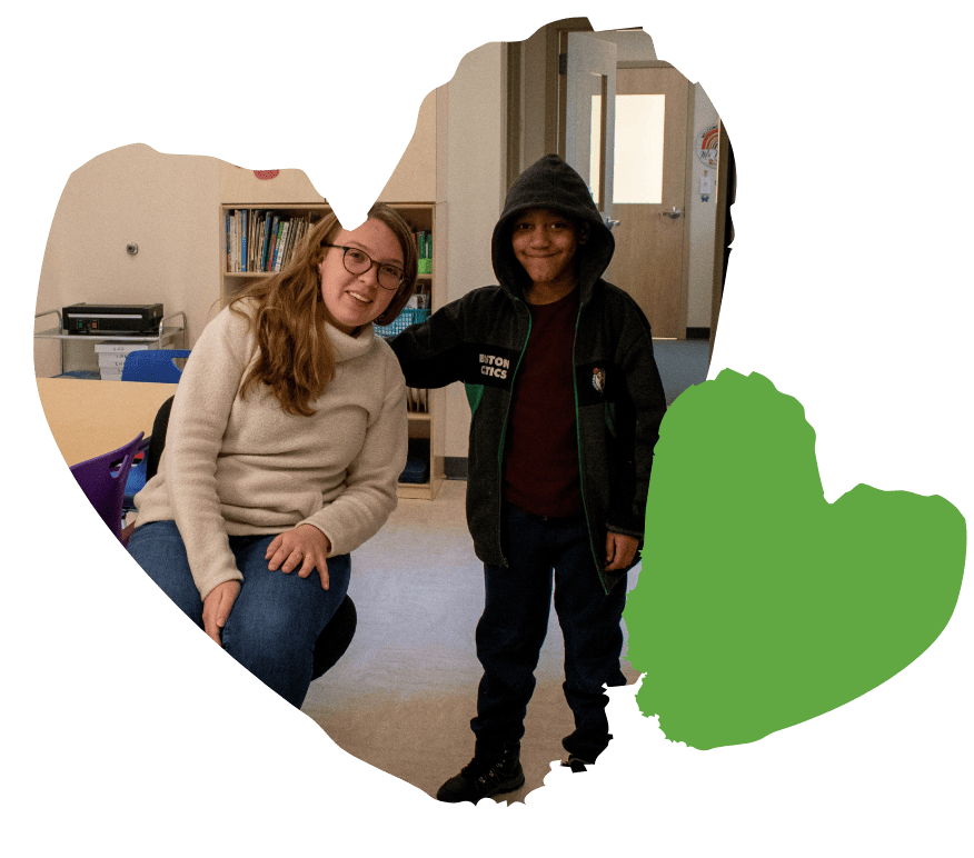 A woman seated next to a smiling child in a classroom, with a green heart outline overlay.