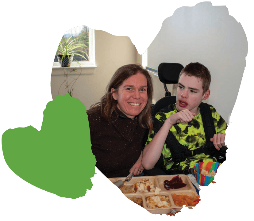 Woman and boy in a wheelchair sitting at a table with food, both smiling. Heart shape overlay and plant in the background.