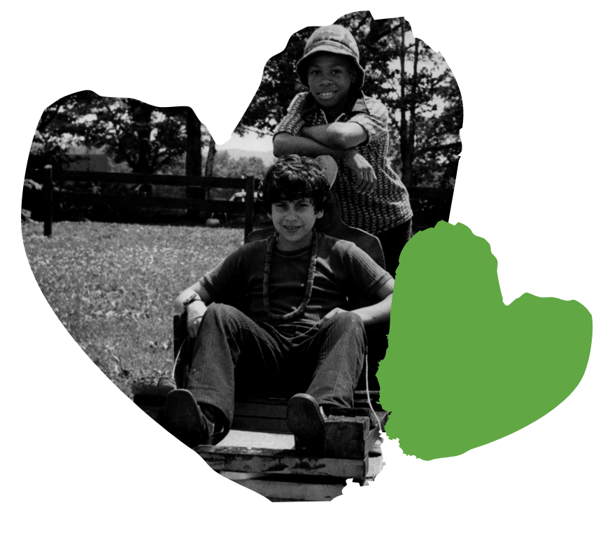 Two children smiling; one sits on a cart, the other leans on it. Theyre outdoors with a green heart shape overlay.
