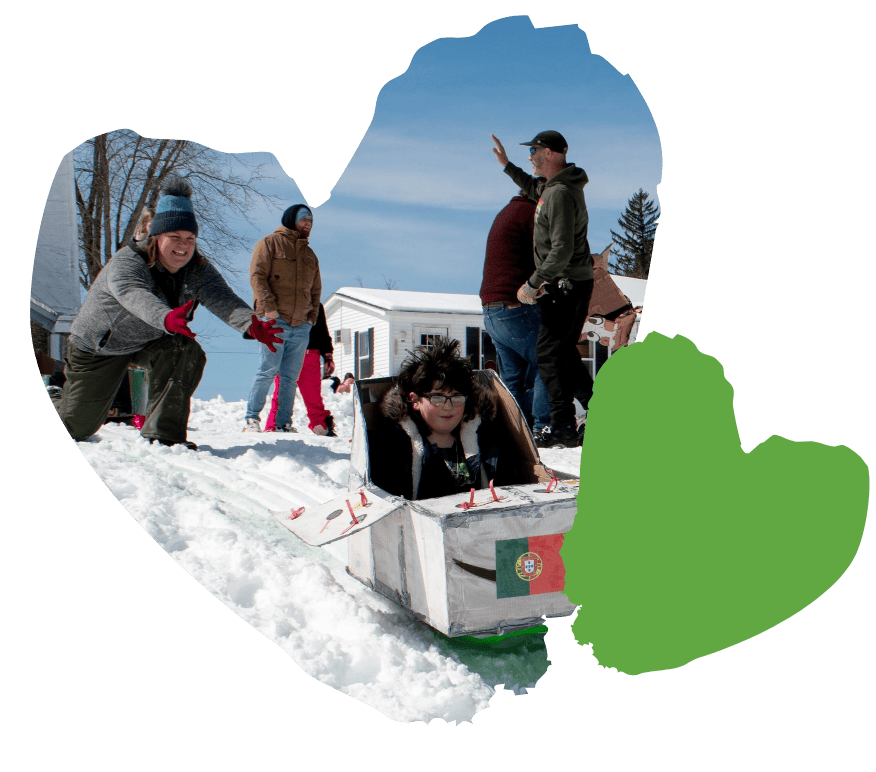 A child rides down a snowy hill in a homemade sled made from cardboard with a Portuguese flag on it. An adult in a blue jacket kneels beside the sled. Other people stand in the background, some wearing coats and hats.