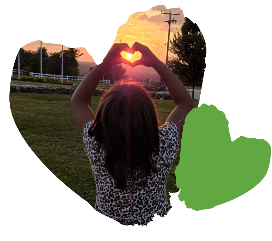 Child forming a heart with hands against a sunset backdrop, surrounded by trees and a fence.