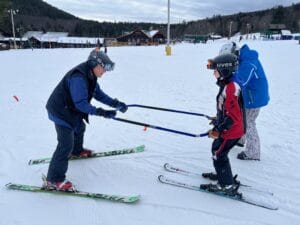 An adult teaching a child to ski using a ski bar on a snowy slope, with another person standing nearby.
