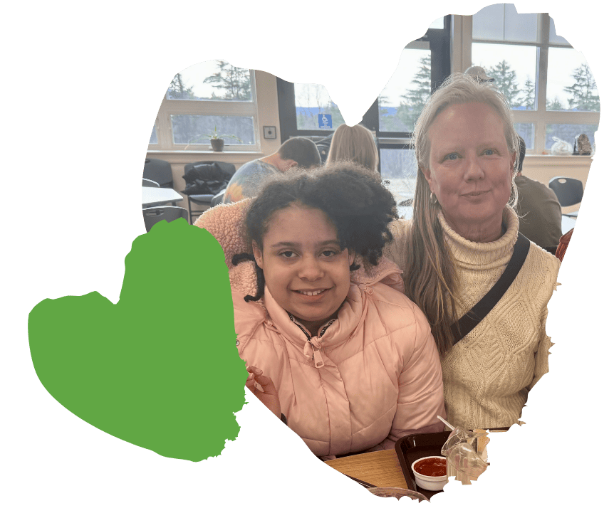 A young girl in a pink jacket sits next to her foster parent, an older woman in a cream sweater. They are inside, surrounded by other people and seated at a table with a lunch tray. A large green heart shape decorates the left side of the image.