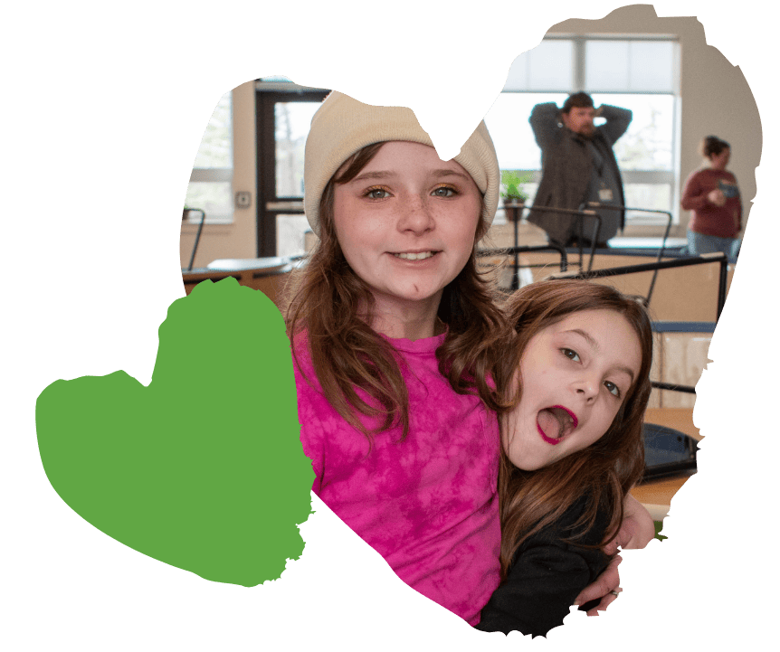 Two smiling girls embrace indoors, with adults and furniture blurred in the background.