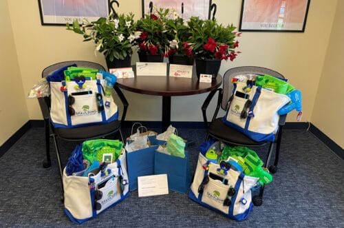 Four gift bags filled with beach toys and essentials on chairs around a table with plants and cards. Four gift bags filled with beach toys and essentials on chairs around a table with plants and cards.