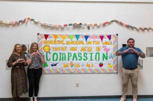 Four people stand in front of a compassion-themed bulletin board, making heart signs with their hands. Four people stand in front of a compassion-themed bulletin board, making heart signs with their hands.
